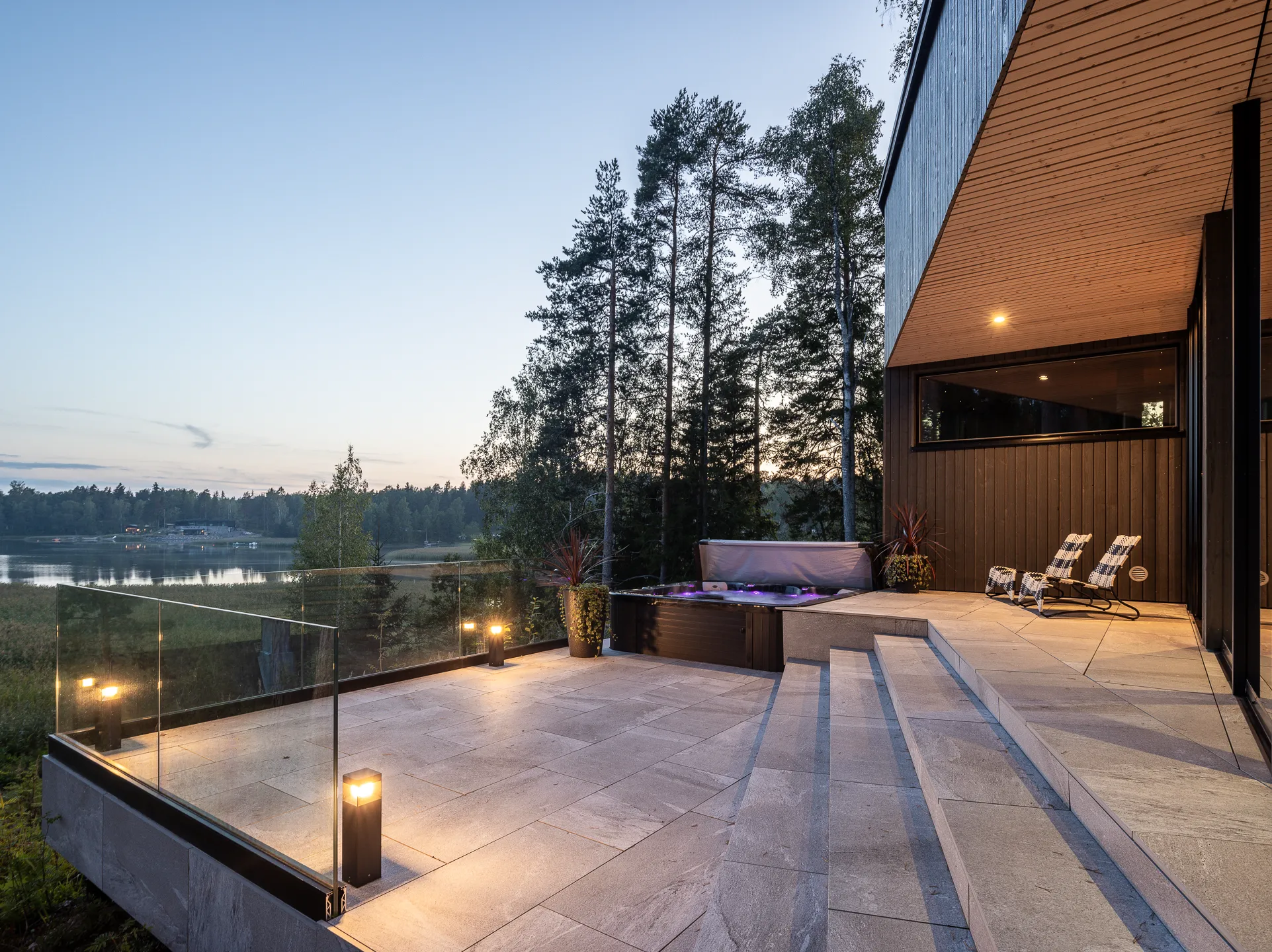 Moderni terassi järvinäköalalla, poreallas, ulkokalusteet ja tunnelmallinen valaistus iltahämärässä.