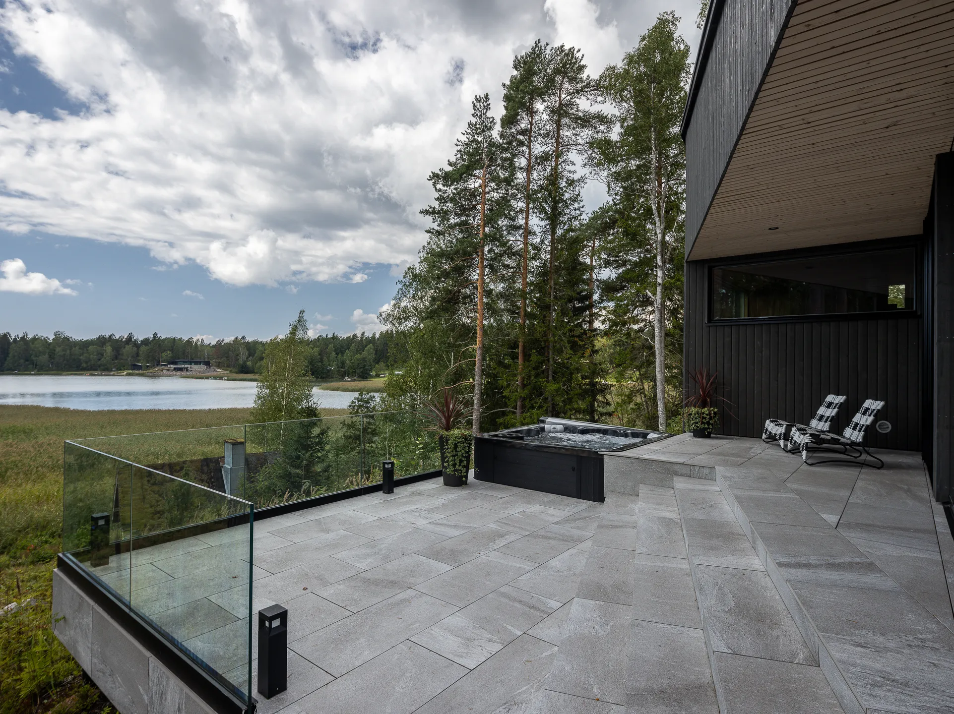 Ulkoterassi järvinäköalalla, jossa on lasikaide, poreallas ja kaksi lepotuolia. Harmaa kivilaatoitus ja tumma puuverhous.