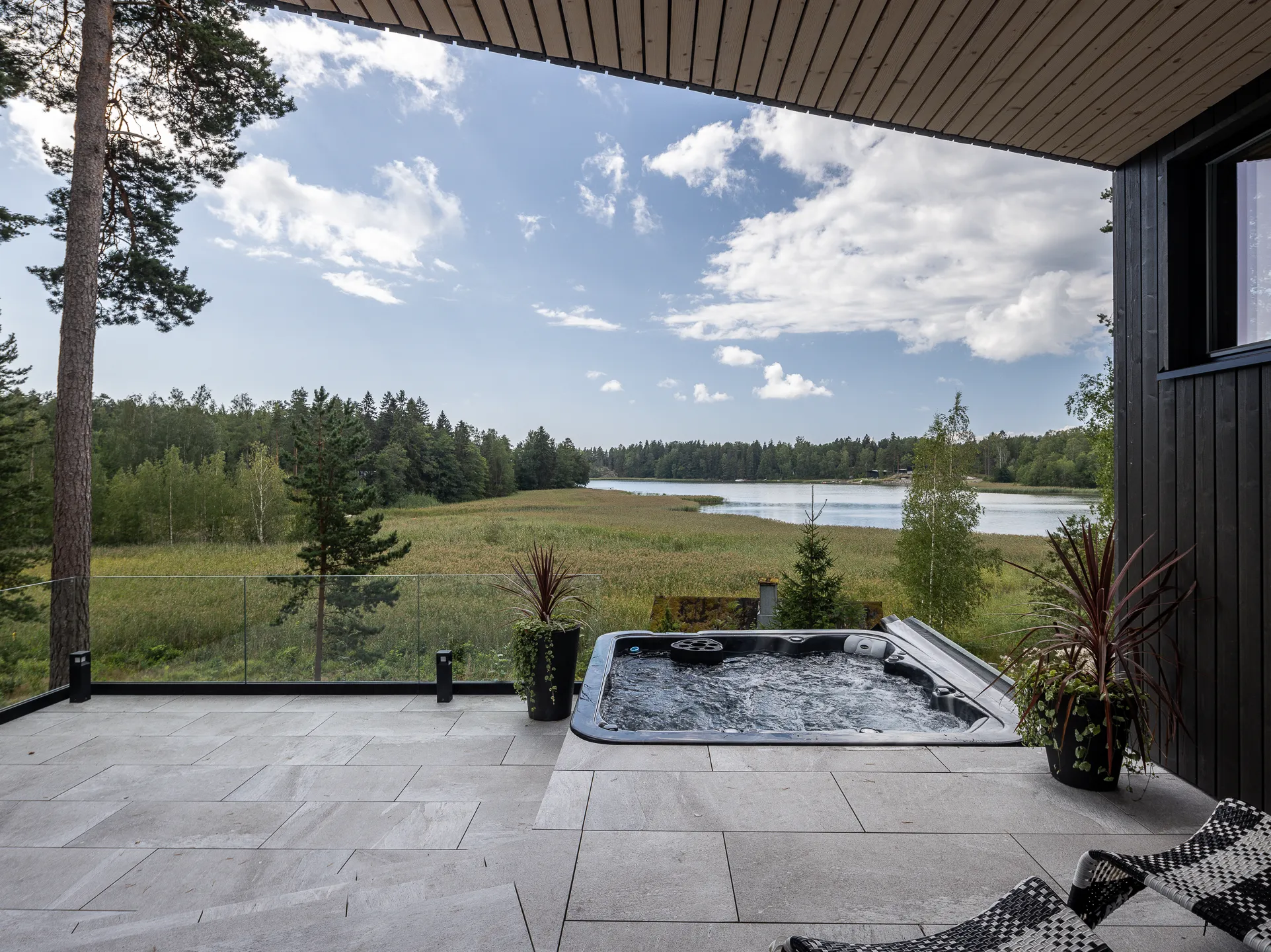 Terassi porealtaalla järvinäköalalla, jossa on harmaat laatat, musta poreallas, kaksi kukkaruukkua ja näkymä järvelle ja metsään aurinkoisena päivänä.