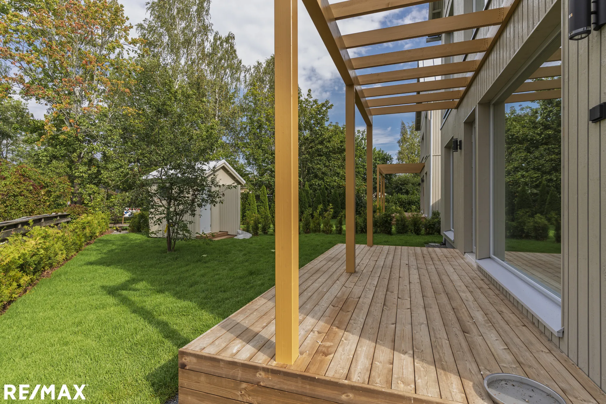 Ulkoterassi puupylväillä, vihreä nurmikko ja pieni puutarhavaja aurinkoisessa säässä.