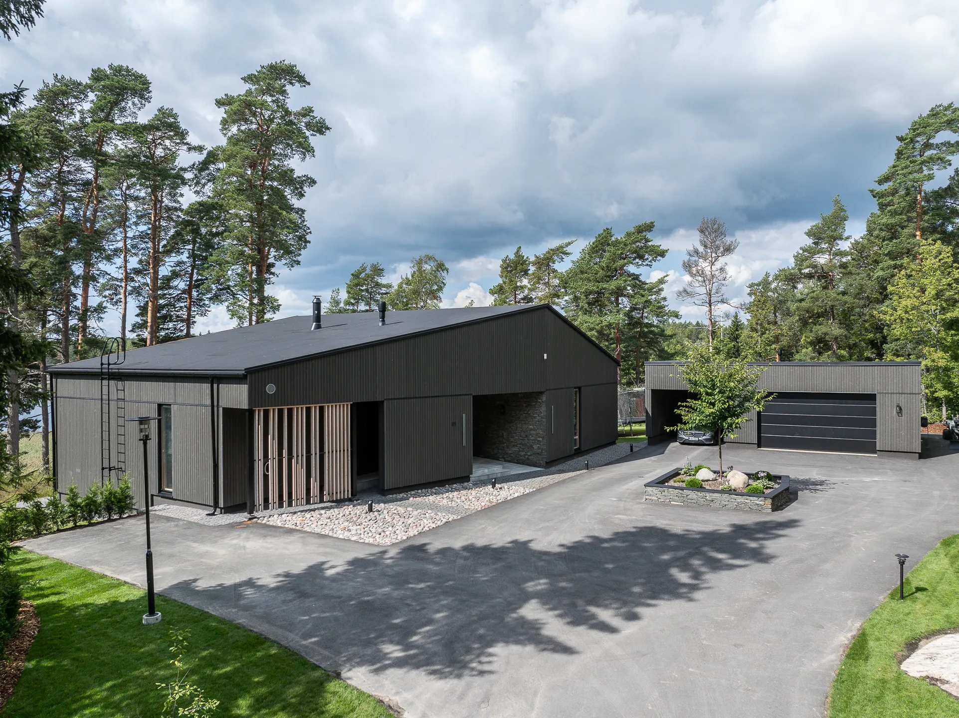 Moderni, tumma puutalo, jossa on harmaa kivipiha ja vihreä nurmikko, valaistus on luonnollinen ja pilvinen.