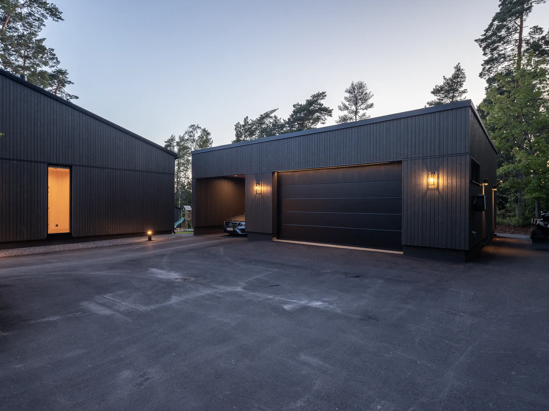 Moderni, tumma puutalo, jossa on autotalli ja valaistu piha-alue iltavalaistuksessa.