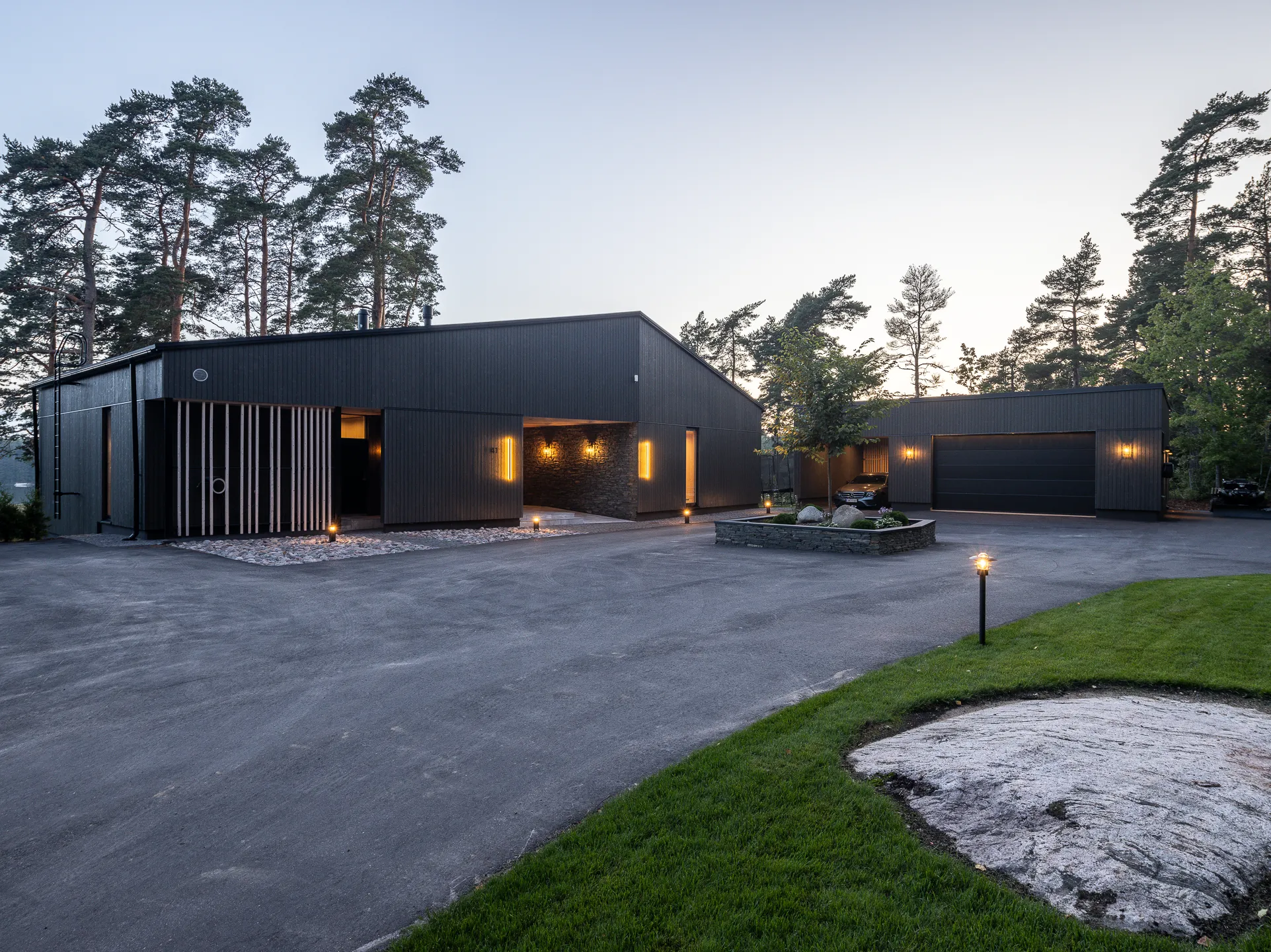 Moderni, tumma puutalo, jossa on valaistu ajotie, kivipuutarha ja nurmikko, iltavalaistuksessa.