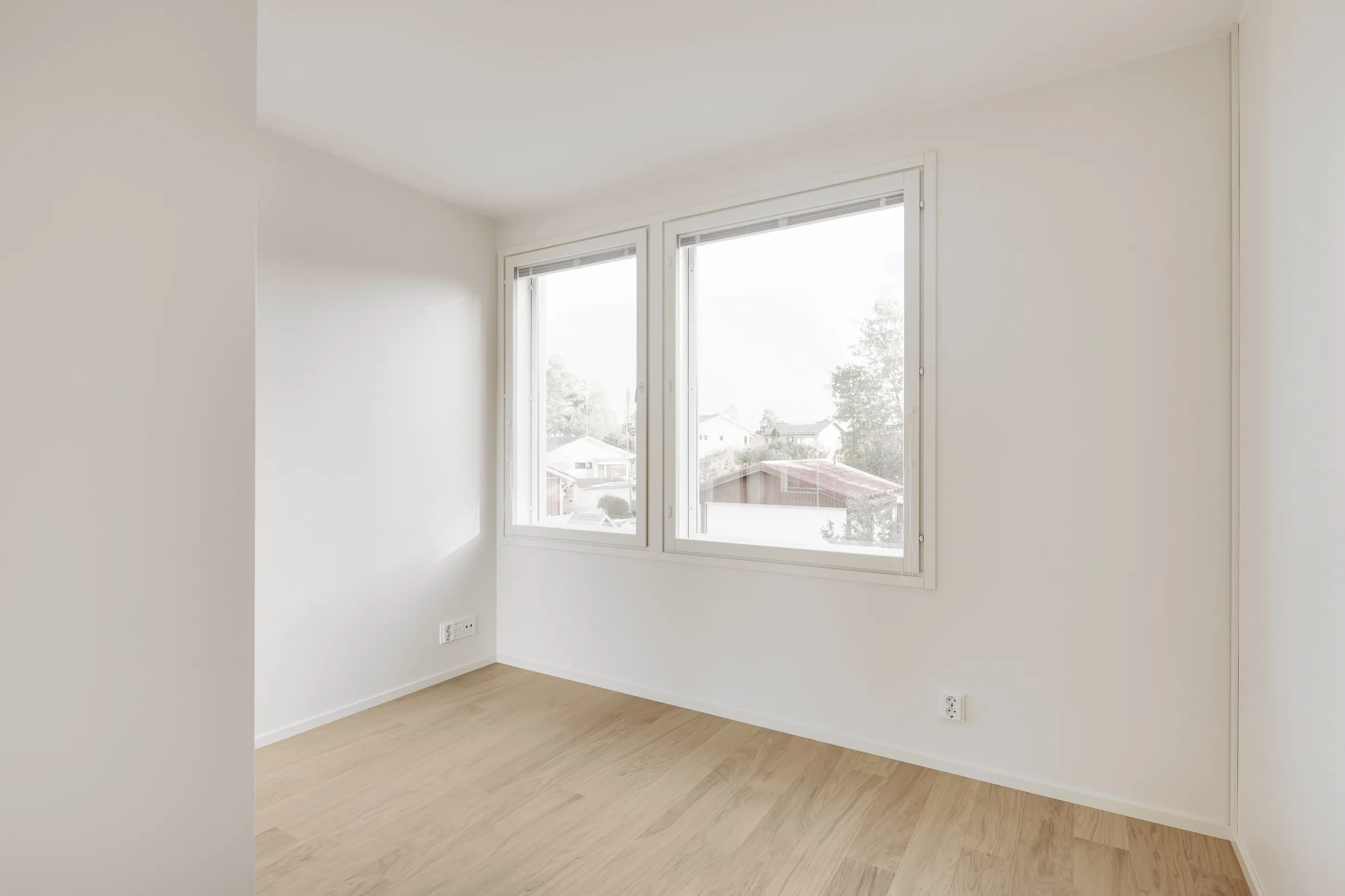 Vaalea, tyhjä huone, jossa on puulattia, kaksi ikkunaa ja valkoiset seinät.