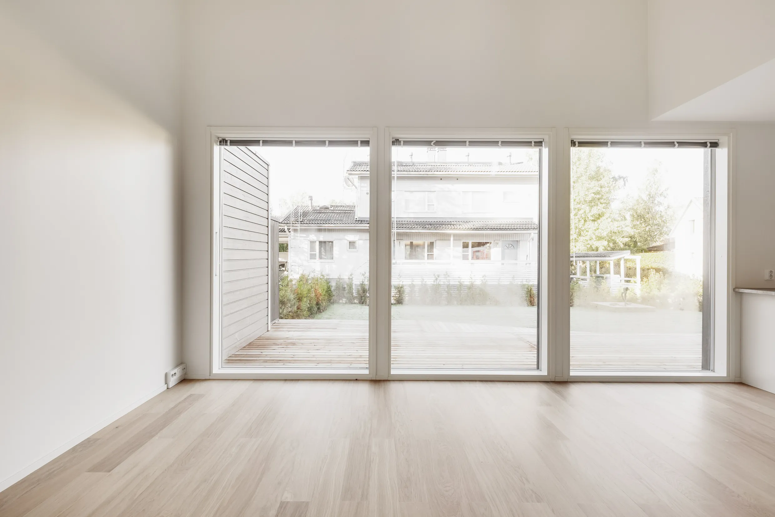 Vaalea, tyhjä huone, jossa on suuret ikkunat, jotka avautuvat terassille ja naapuritaloon, sekä vaaleat puulattiat.