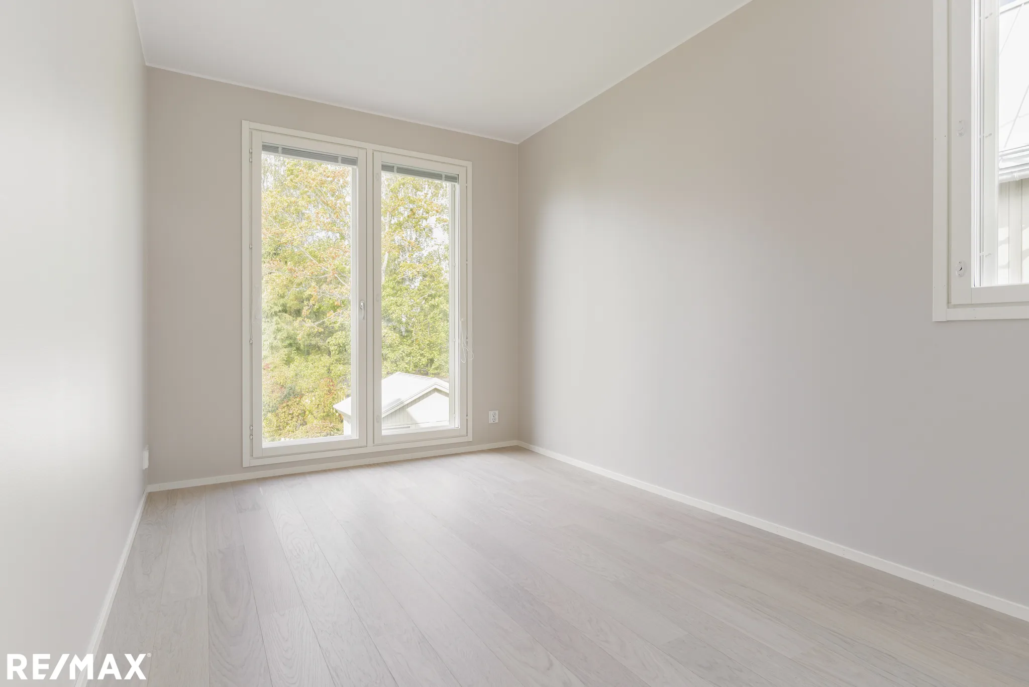 Vaalea, tyhjä huone, jossa on suuret ikkunat ja vaaleat puulattiat. Seinät ovat vaaleanharmaat ja huone on valoisa.