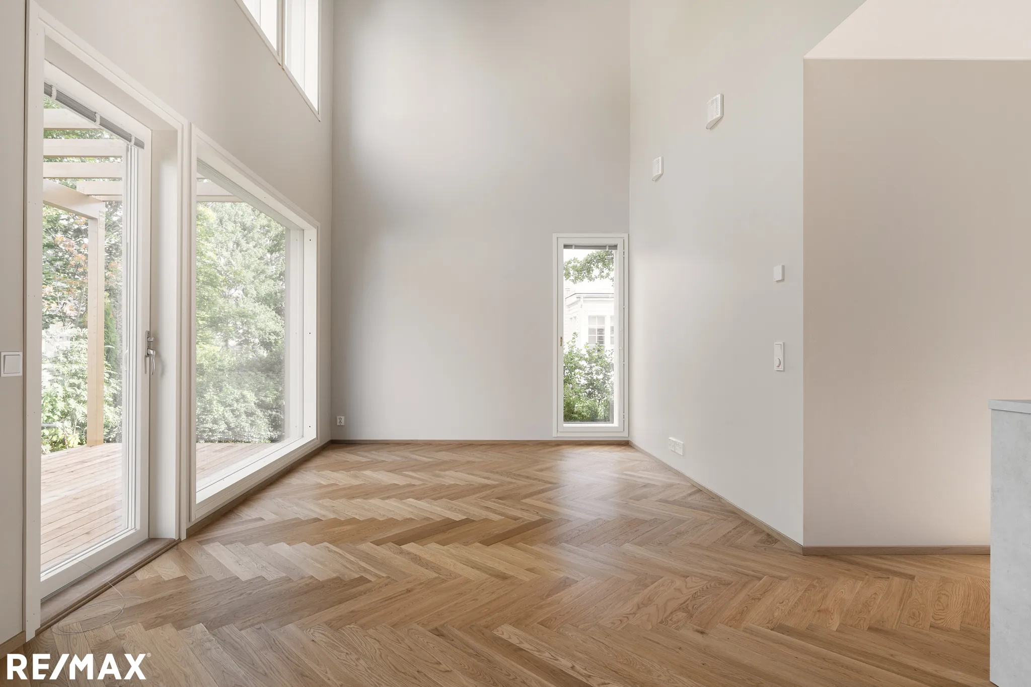 Vaalea, tyhjä huone, jossa on parkettilattia, suuret ikkunat ja korkea katto. RE/MAX-logo alhaalla vasemmalla.
