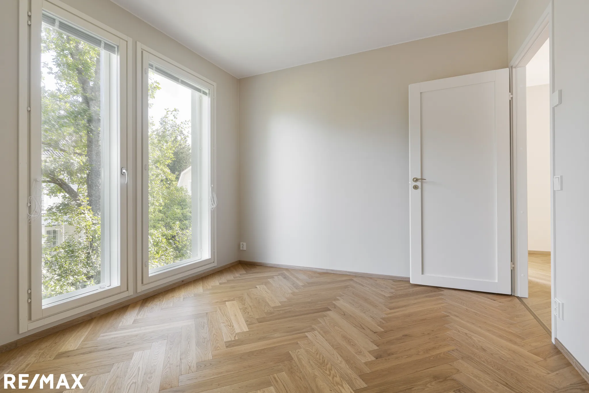 Vaalea huone, jossa on parkettilattia, kaksi ikkunaa ja avoin valkoinen ovi.