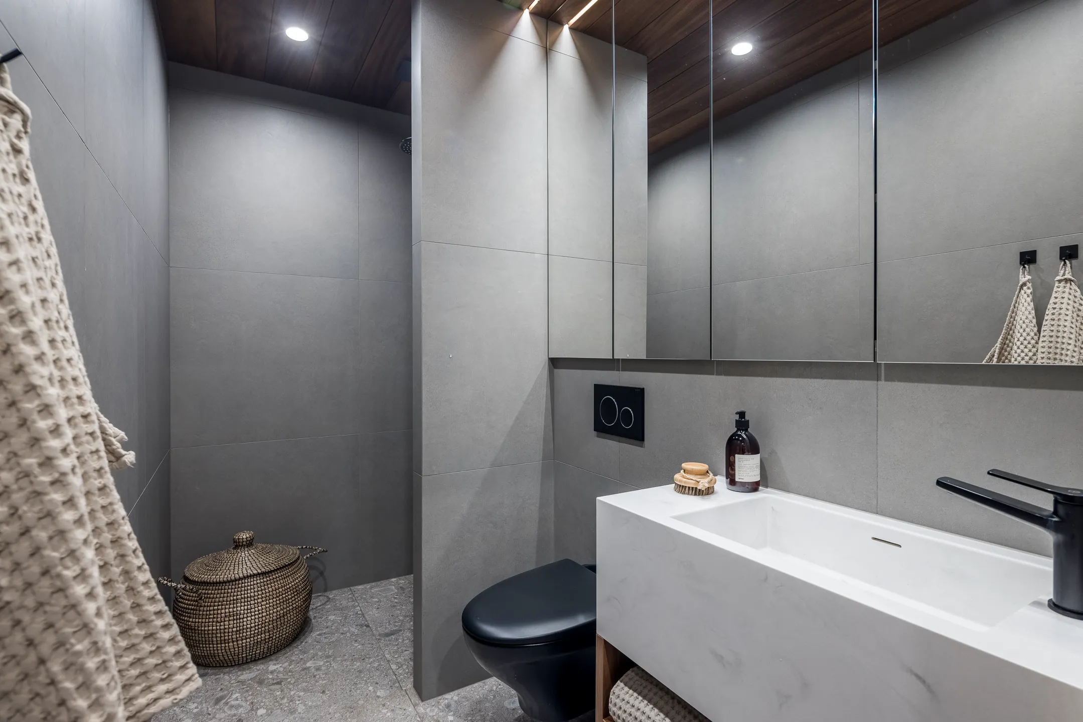 Moderni kylpyhuone harmaalla laatoituksella, mustalla WC:llä ja valkoisella pesualtaalla. Puinen katto ja peilikaappi lisäävät lämpöä.