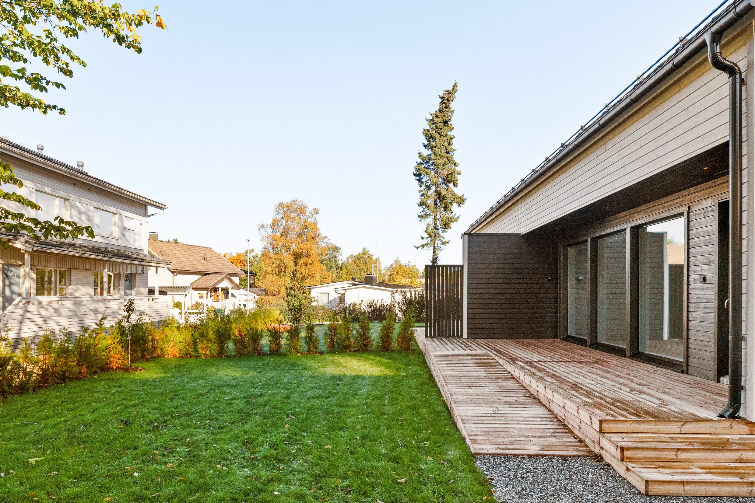 Valoisa kuva ulkotilasta, jossa on nurmikko, puutalo ja puinen terassi. Terassi on ruskea ja siinä on liukuovet. Taustalla näkyy muita taloja ja puita syksyn väreissä.
