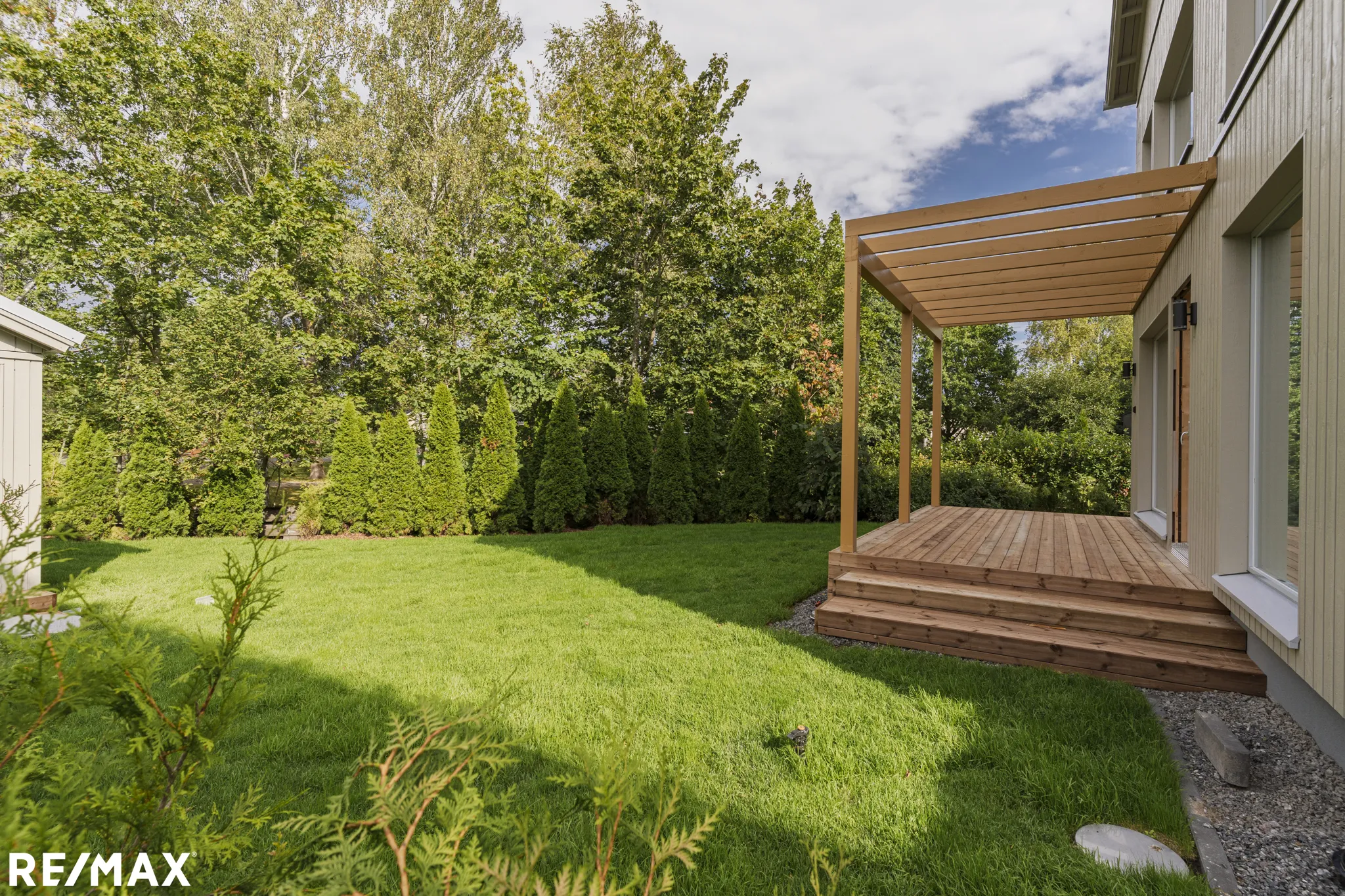 Ulkona on nurmikko, puita ja puinen terassi, jossa on pergola. RE/MAX-logo näkyy alareunassa.