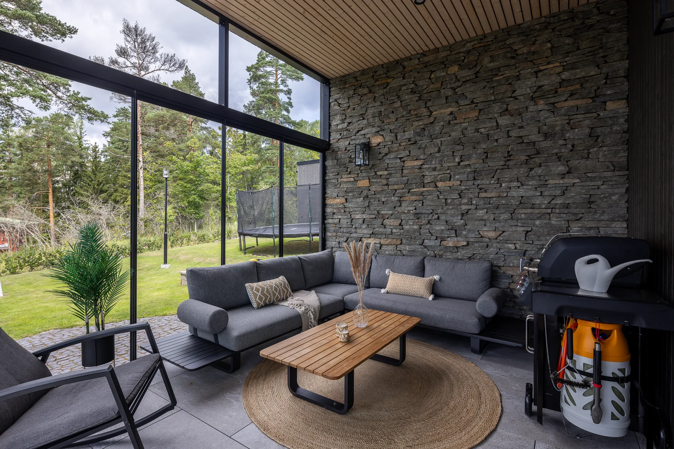 Valoisassa ulkotilassa on harmaa kulmasohva, puinen sohvapöytä, pyöreä matto, grilli ja viherkasveja. Suuret ikkunat avautuvat vehreään maisemaan.