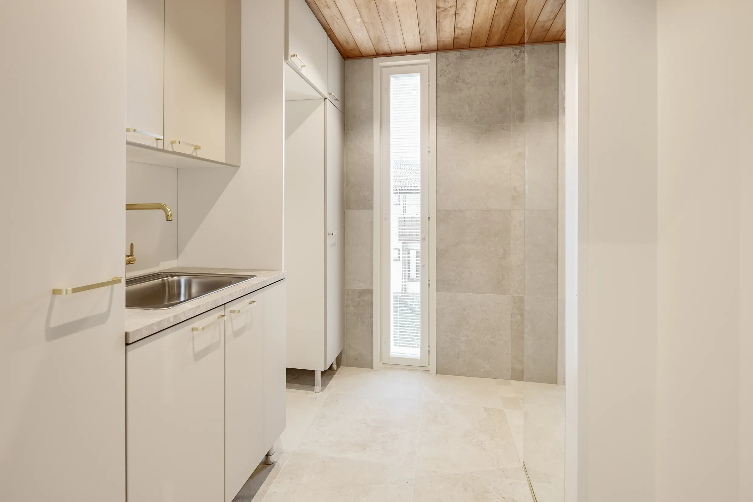 Vaalean huoneen sisustus, jossa on puukatto, valkoiset kaapit, teräksinen pesuallas ja ikkuna, jossa on sälekaihtimet.