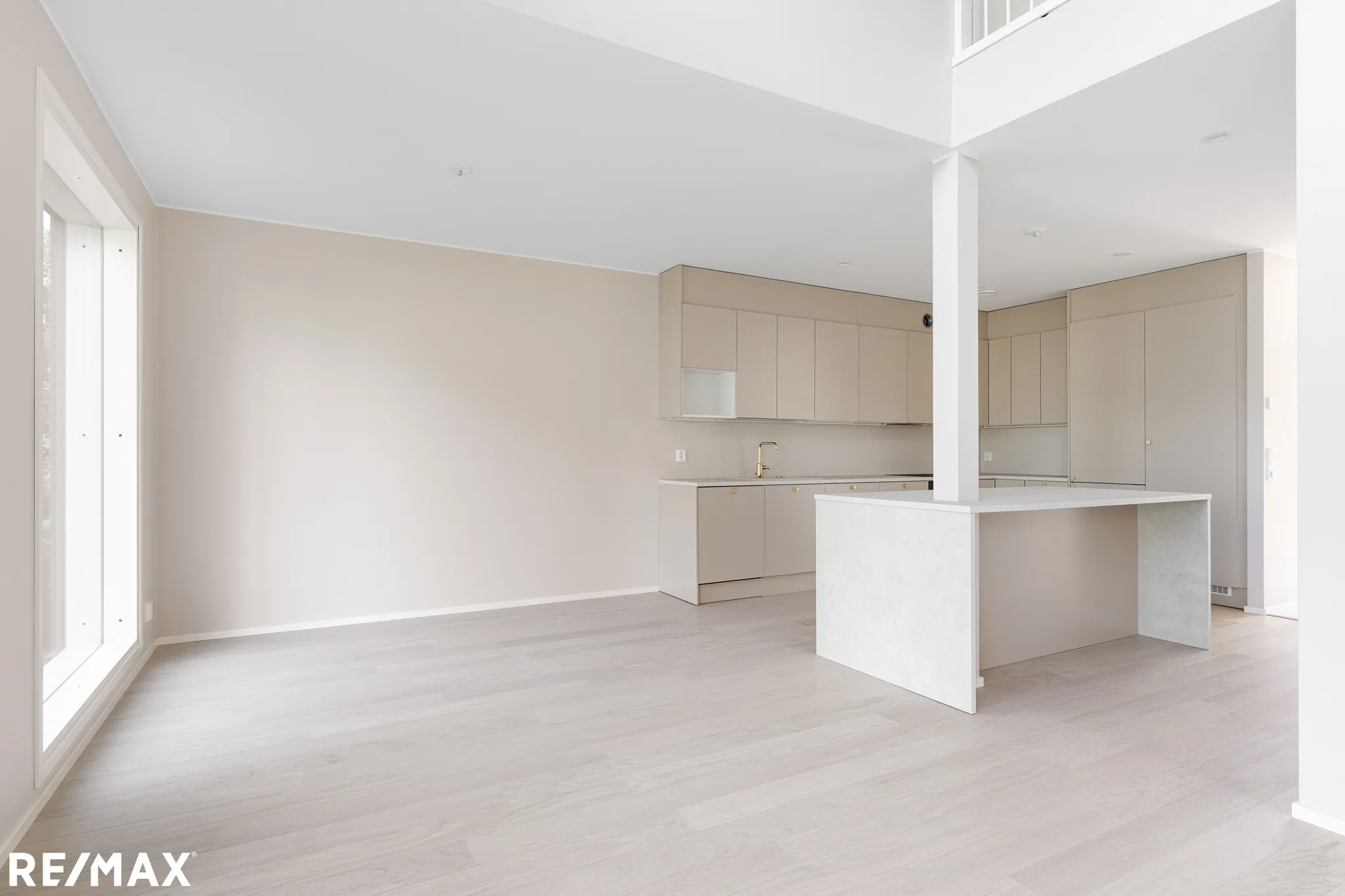 Vaalean sävyinen, avara keittiö-olohuone, jossa on valkoiset seinät, vaaleat puulattiat ja moderni keittiö saarekkeella. Suuret ikkunat tuovat runsaasti luonnonvaloa.