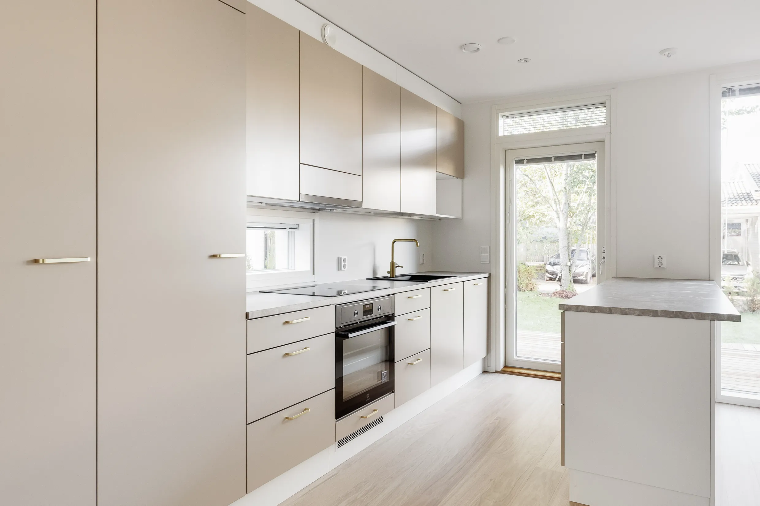 Vaalean keittiön sisustus, jossa on vaaleat kaapit, kultaiset vetimet, musta pesuallas ja uuni. Ikkunasta näkyy ulos.