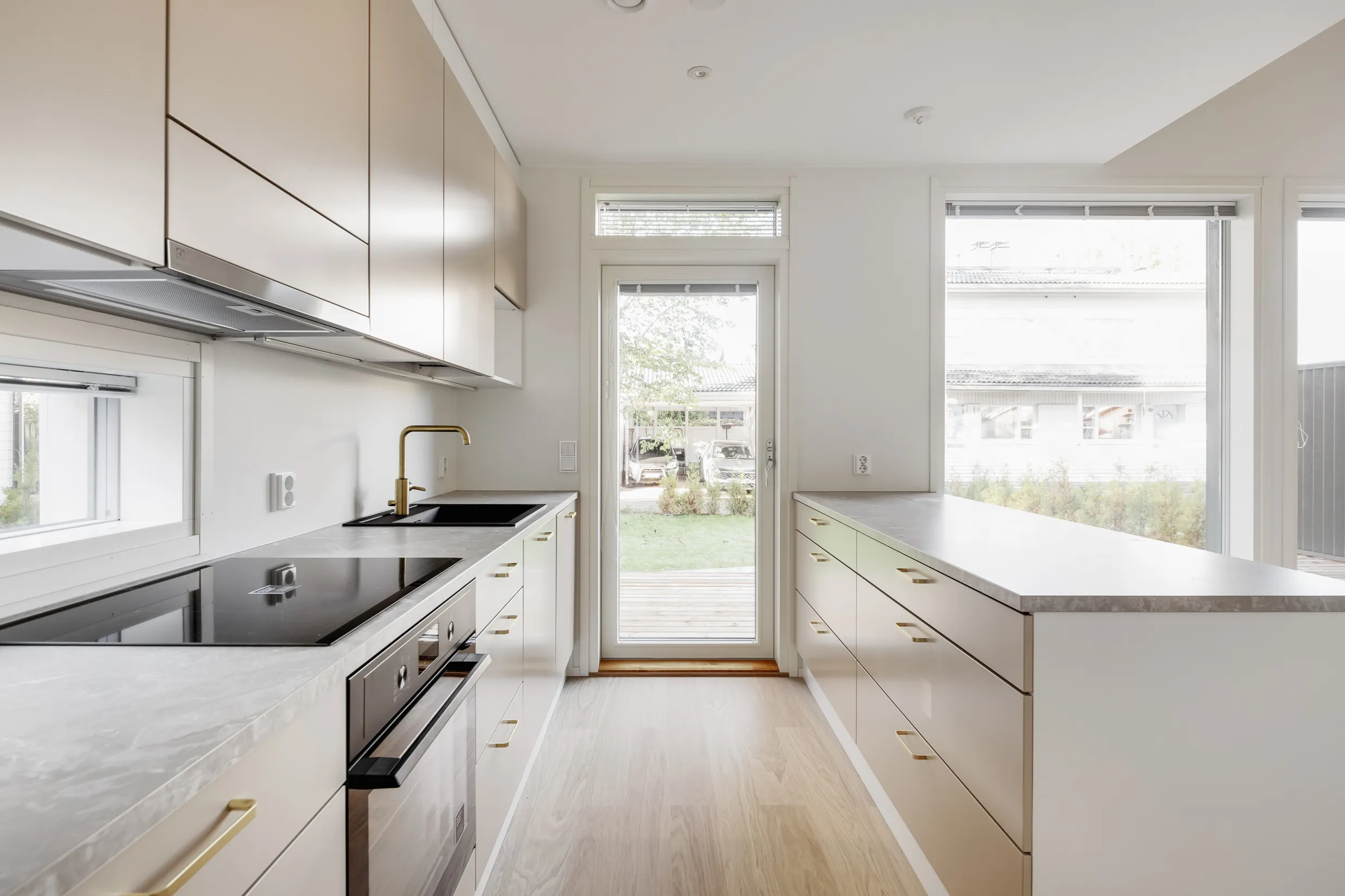 Vaaleassa keittiössä on vaaleat kaapit, marmorikuvioiset työtasot, kultainen hana ja musta liesi. Ikkunoista näkyy ulos.