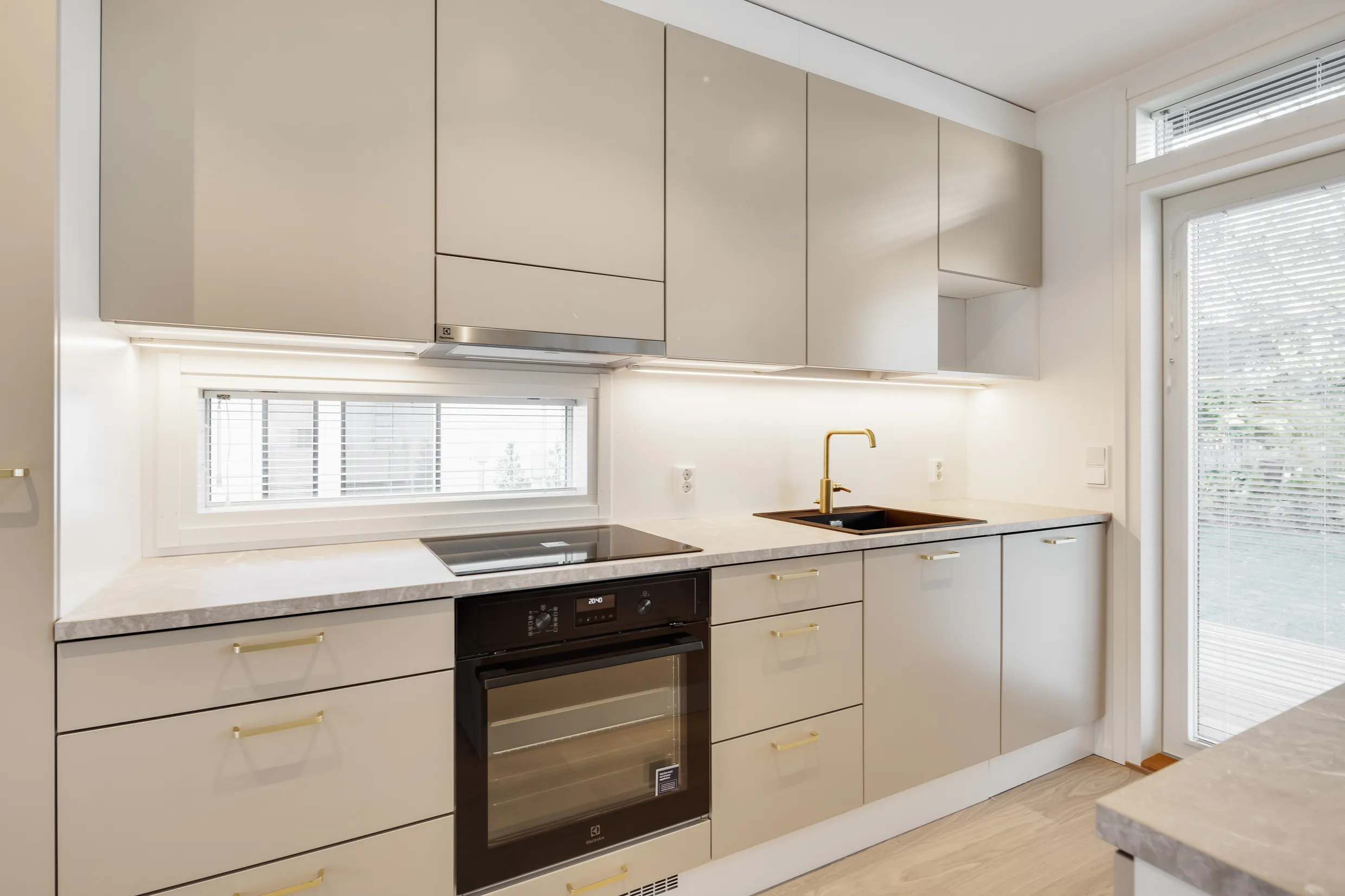 Vaalean keittiönäkymä, jossa on uuni, liesi, tiskiallas ja kultainen hana, sekä yläkaapit ja ikkuna.