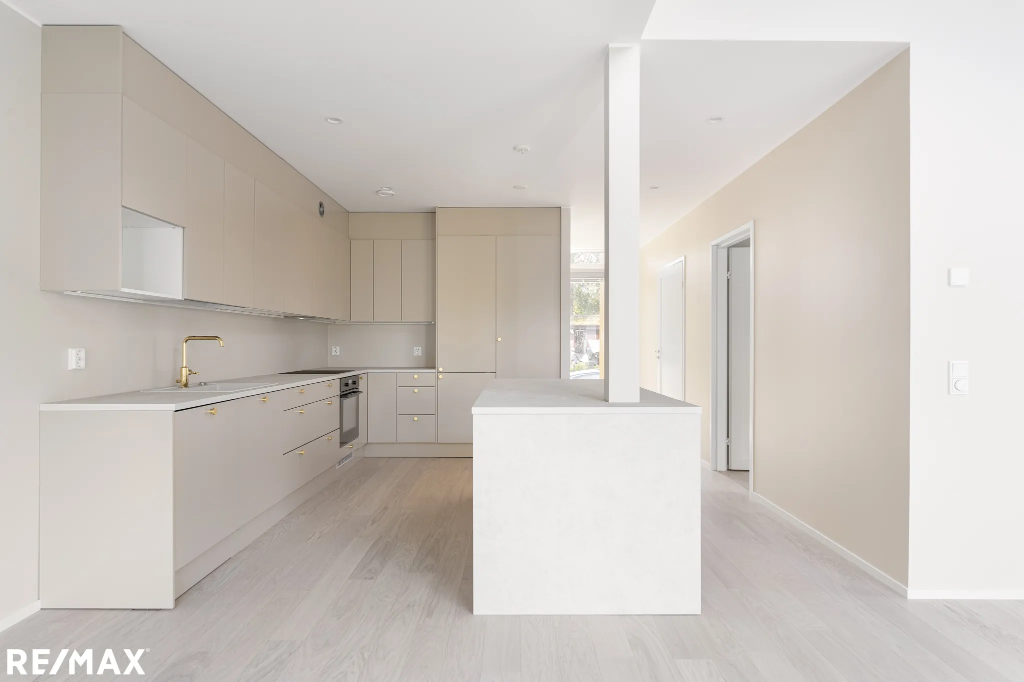 Vaalean keittiön ja olohuoneen sisustus, jossa on vaaleat kaapit, kultainen hana ja valoisa tunnelma.