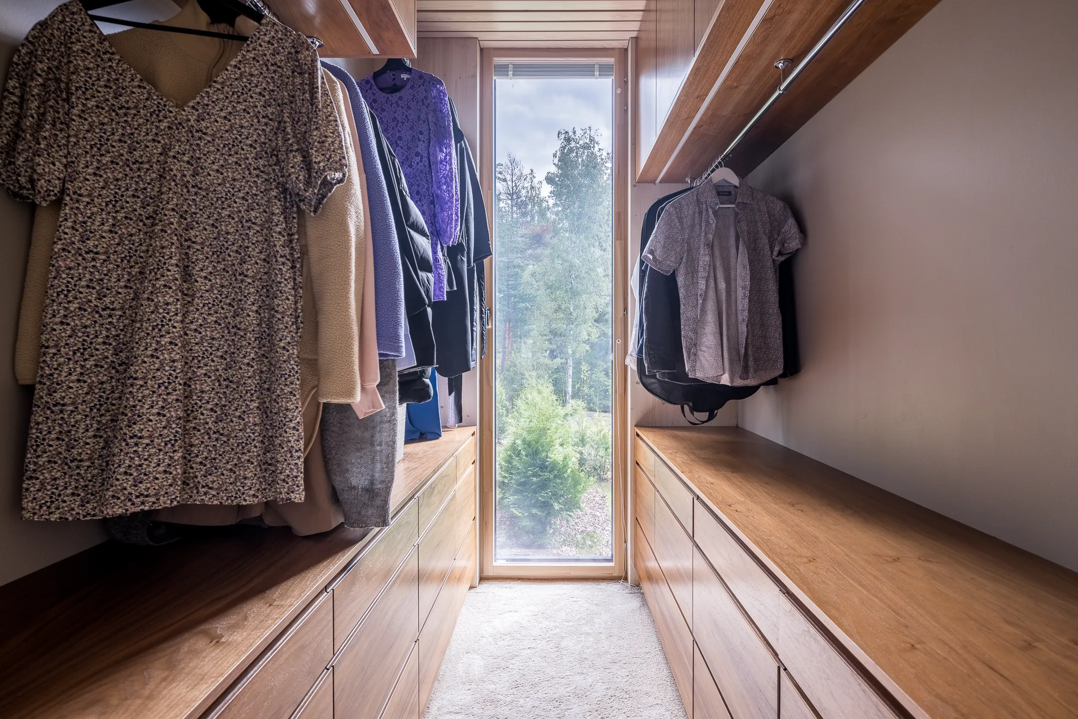 Vaatehuone, jossa on puulaatikot, vaatteita henkareissa ja ikkuna, josta näkyy ulos. Valoisa ja avara tila.