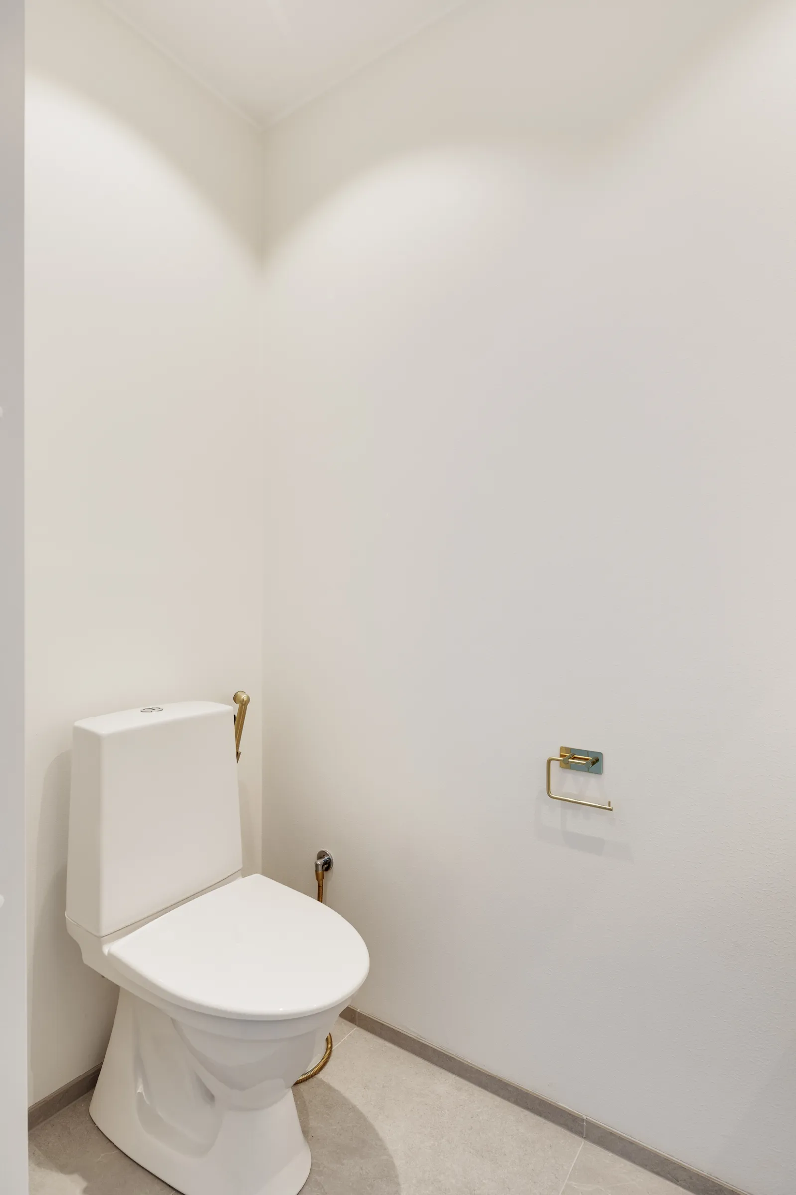 Valkoinen wc-istuin, kultainen paperiteline ja harmaa lattia valoisassa, minimalistisessa kylpyhuoneessa.