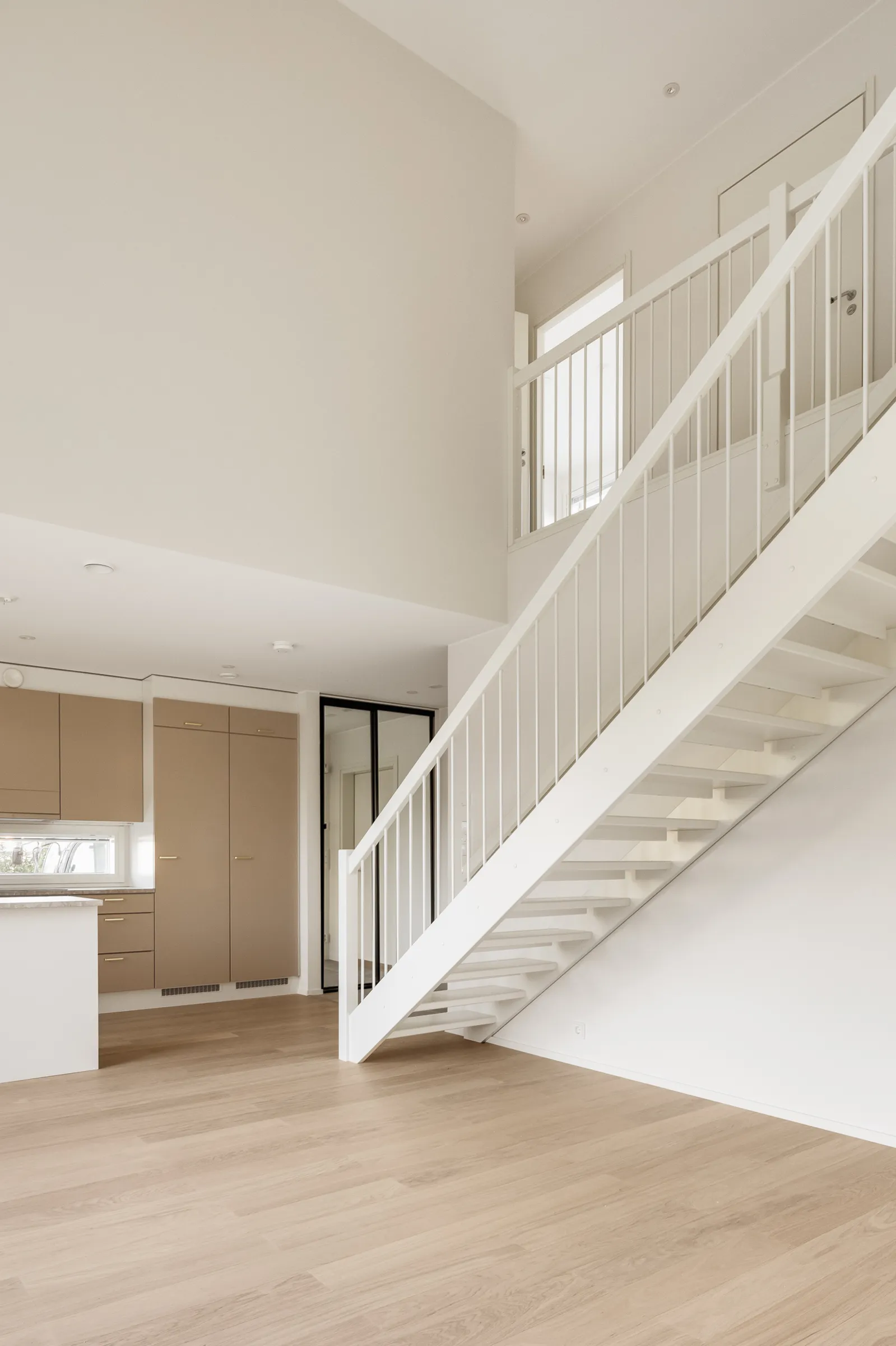 Vaalea, avoin sisätila, jossa on valkoiset portaat, vaaleat puulattiat ja beige keittiö.