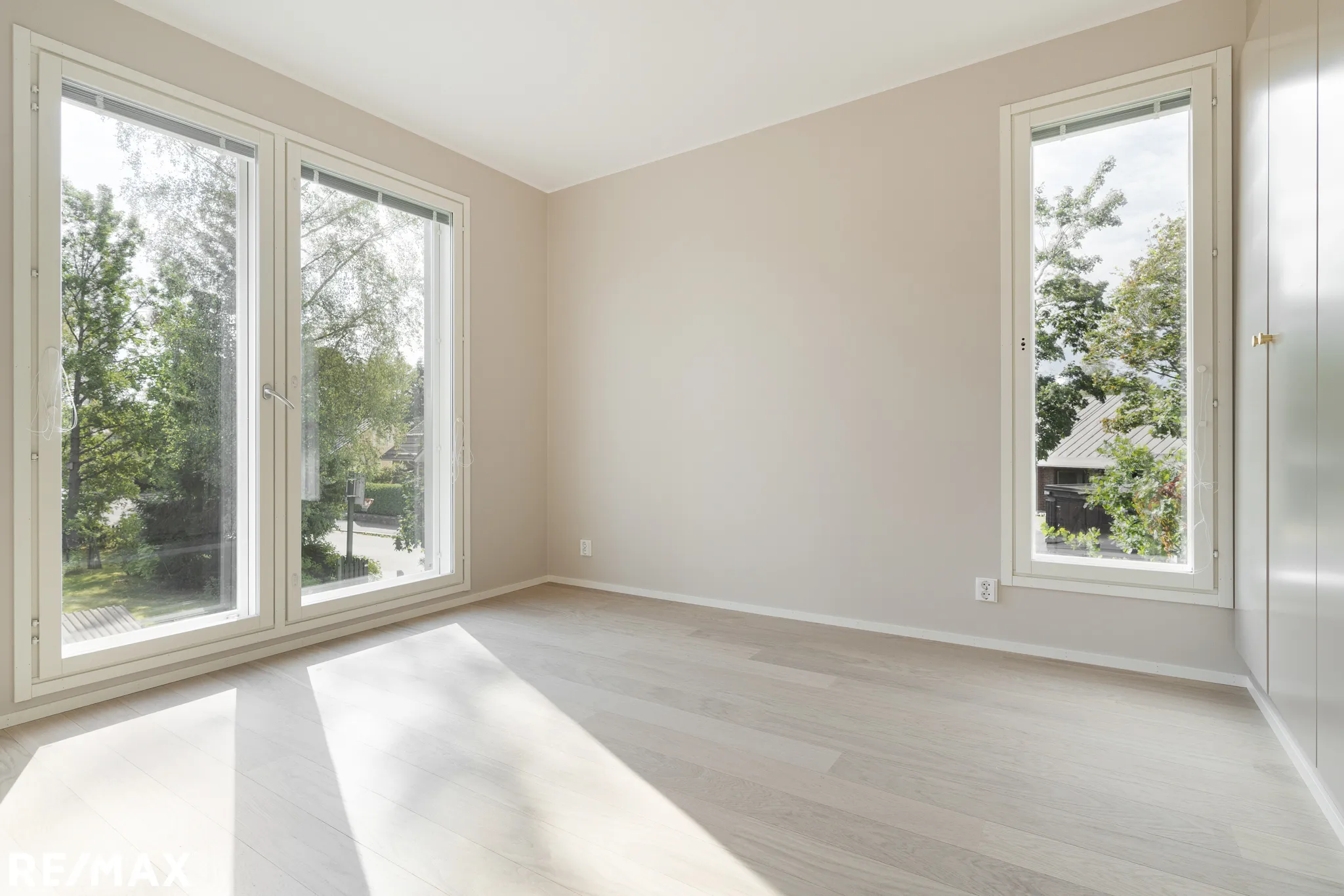 Valoisa huone, jossa on suuret ikkunat, vaaleat seinät ja vaalea puulattia. Auringonvalo lankeaa lattialle.