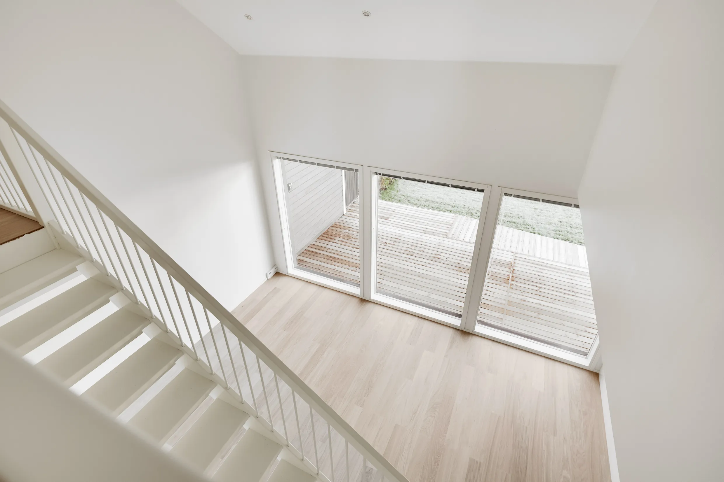 Vaalean huoneen näkymä ylhäältä, jossa valkoiset portaat, suuret ikkunat ja vaalea puulattia.