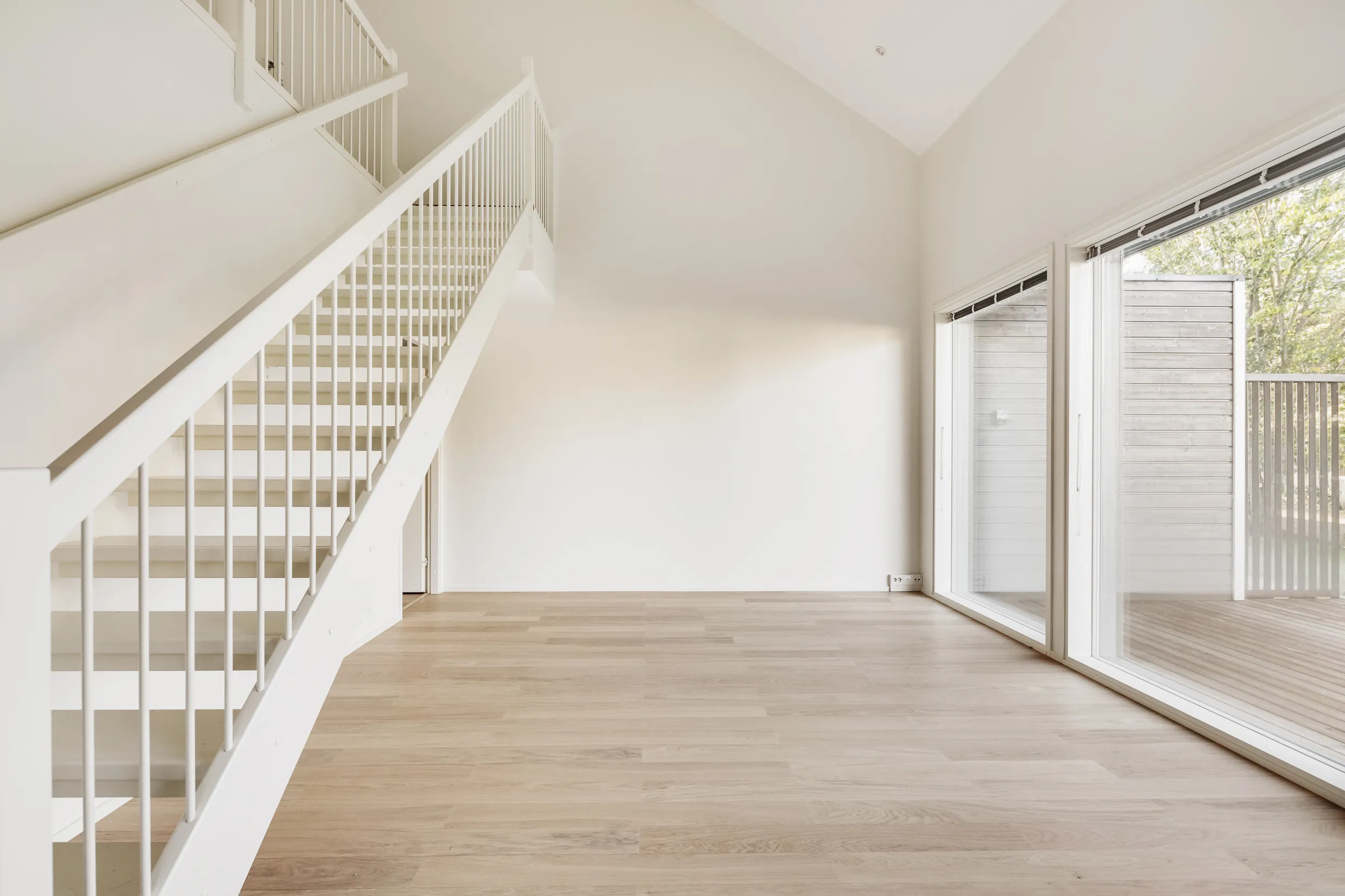 Vaalea, avara huoneisto, jossa valkoiset portaat, suuret ikkunat ja vaalea puulattia.