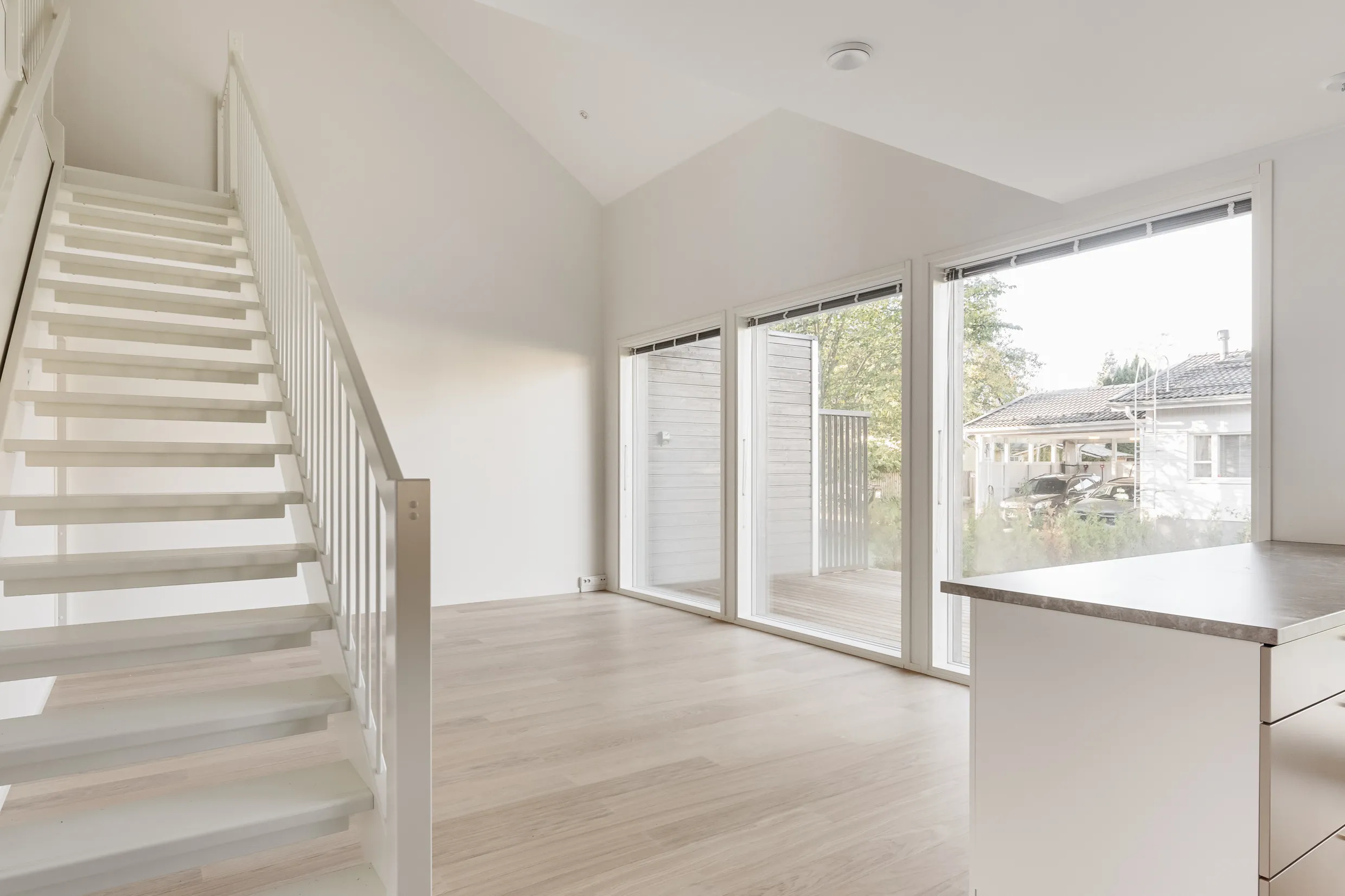 Vaaleassa huoneessa on valkoiset portaat, suuret ikkunat ja vaalea puulattia. Ikkunoista näkyy ulos naapuritalo ja puita.