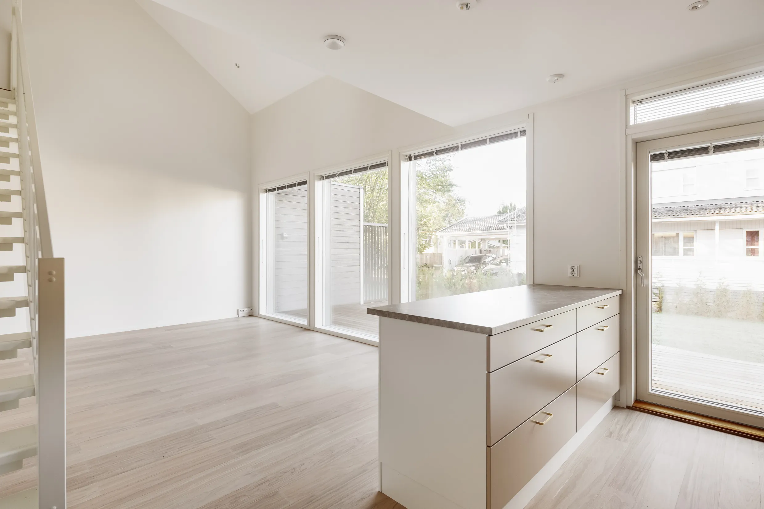 Vaaleassa huoneessa on valkoiset portaat, suuret ikkunat, vaaleat puulattiat ja keittiösaareke, jossa on harmaa työtaso ja kultaiset vetimet.