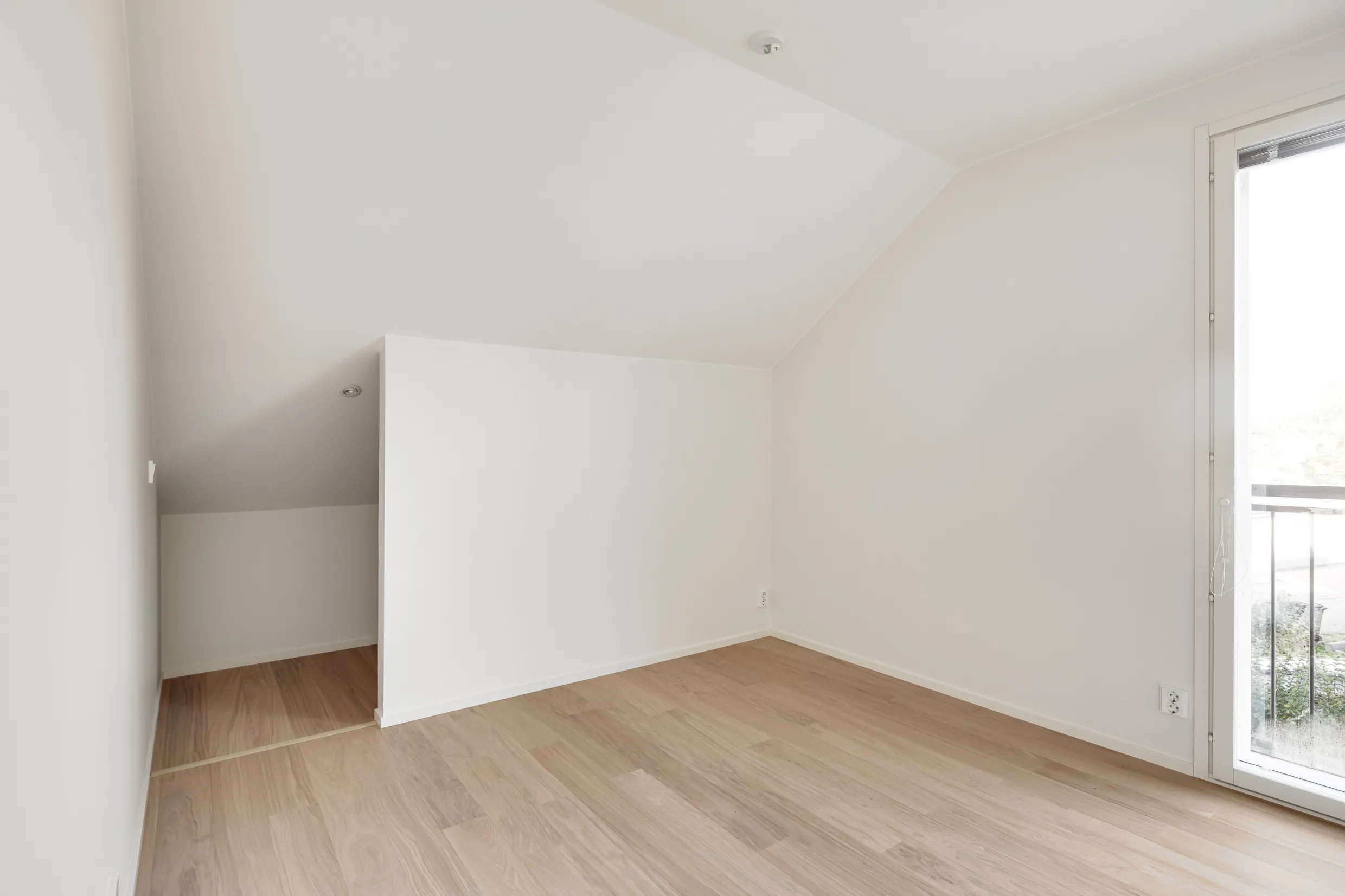 Vaalea huone, jossa on puulattia, valkoiset seinät ja ikkuna parvekkeelle.