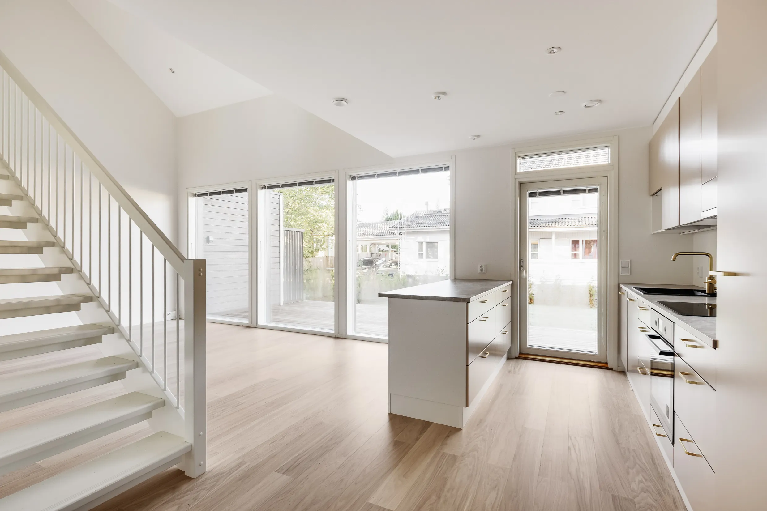 Vaalea, avara huoneisto, jossa valkoiset portaat, suuret ikkunat ja moderni keittiö saarekkeella.