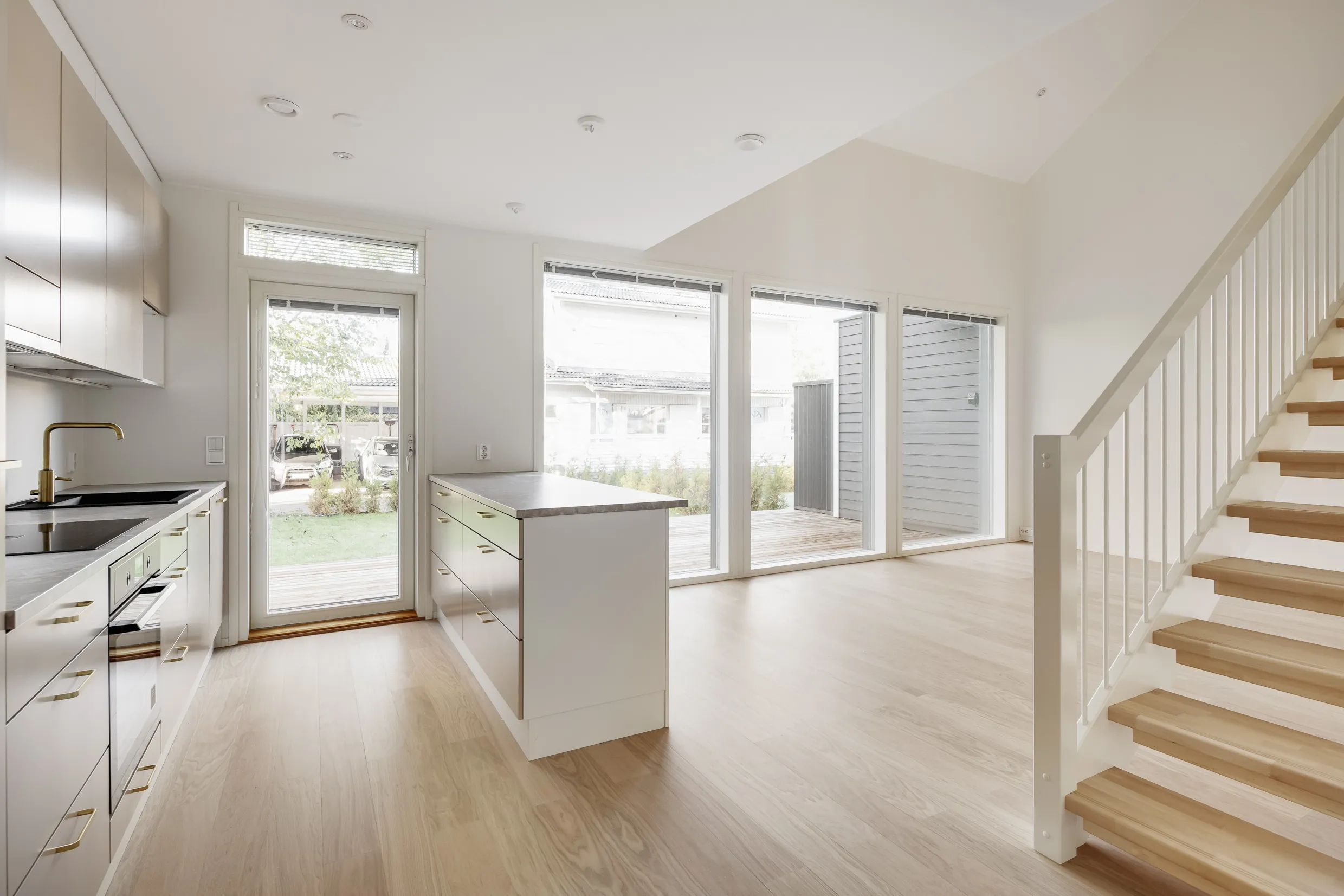 Vaalea keittiö, saareke, suuret ikkunat ja puuportaat valoisassa huoneessa.