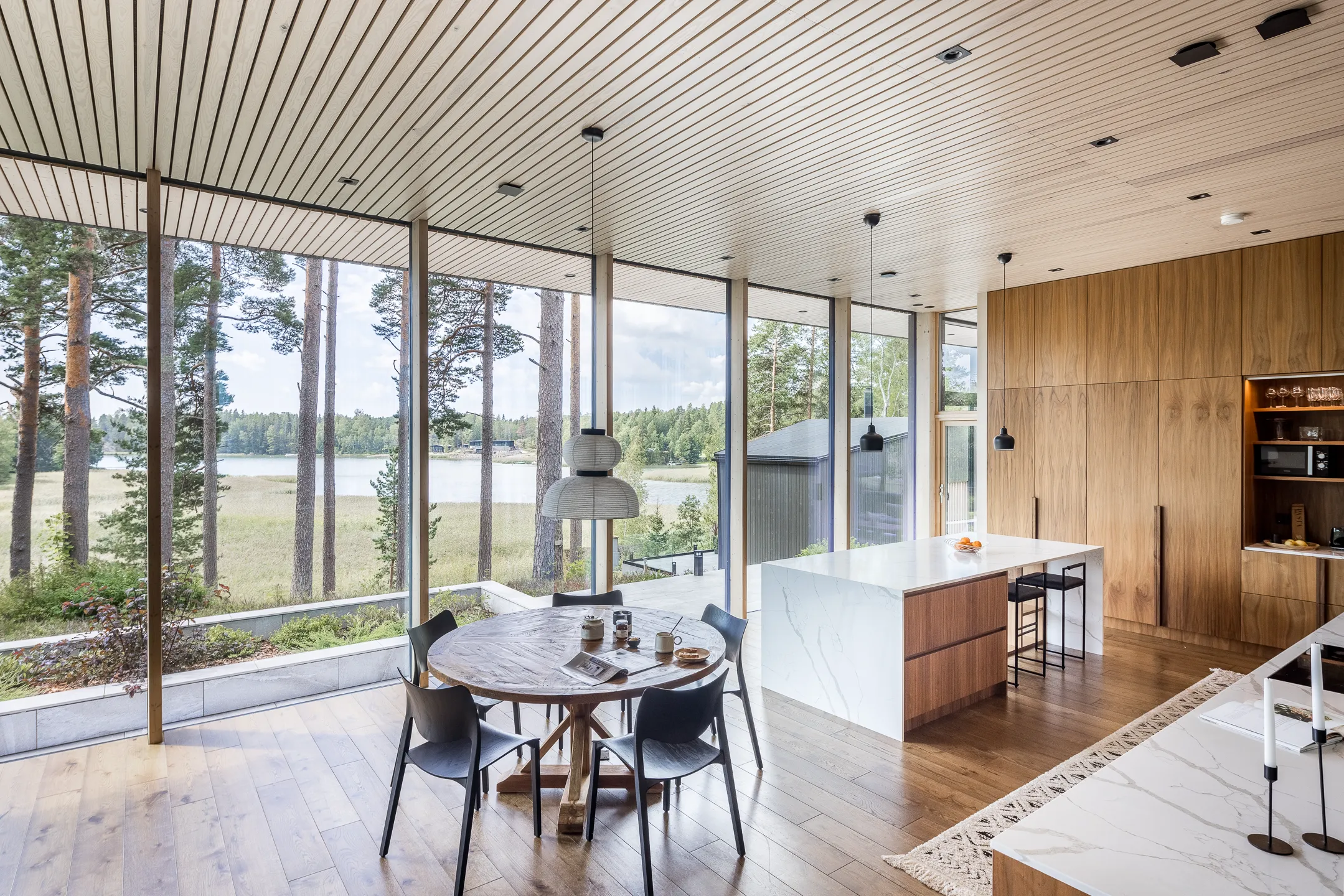 Valoisa ja moderni ruokailutila, jossa on suuret ikkunat, puinen ruokapöytä ja mustat tuolit, sekä avokeittiö, jossa on valkoiset työtasot ja puukaapit.