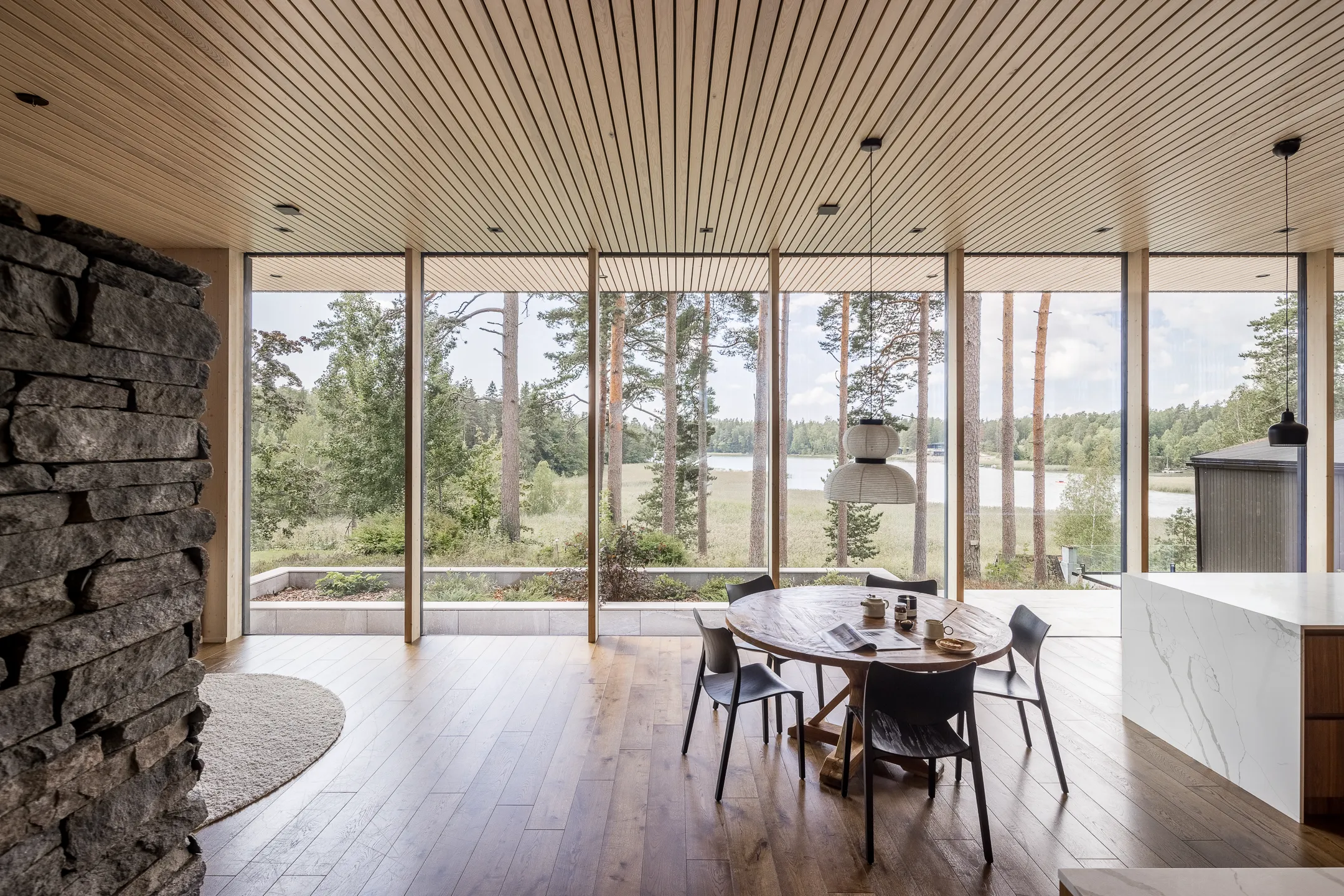 Valoisa ruokailutila, jossa puinen katto, suuret ikkunat, kiviseinä, pyöreä pöytä ja mustat tuolit, luoden rauhallisen ja modernin tunnelman.