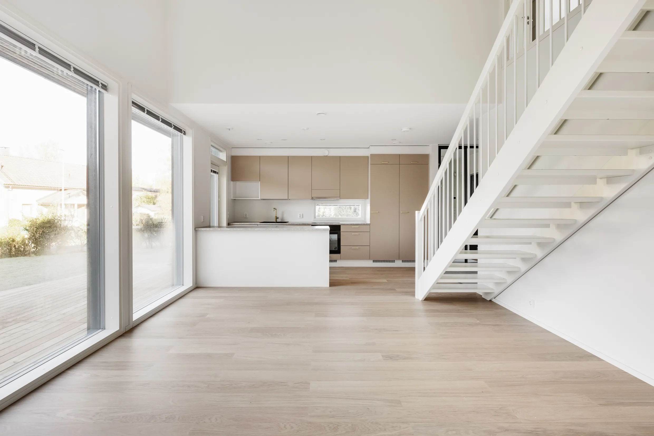 Valoisa ja avara sisätila, jossa on vaaleat puulattiat, valkoiset portaat ja suuret ikkunat, jotka tuovat luonnonvaloa. Keittiössä on beiget kaapit ja valkoinen saareke.