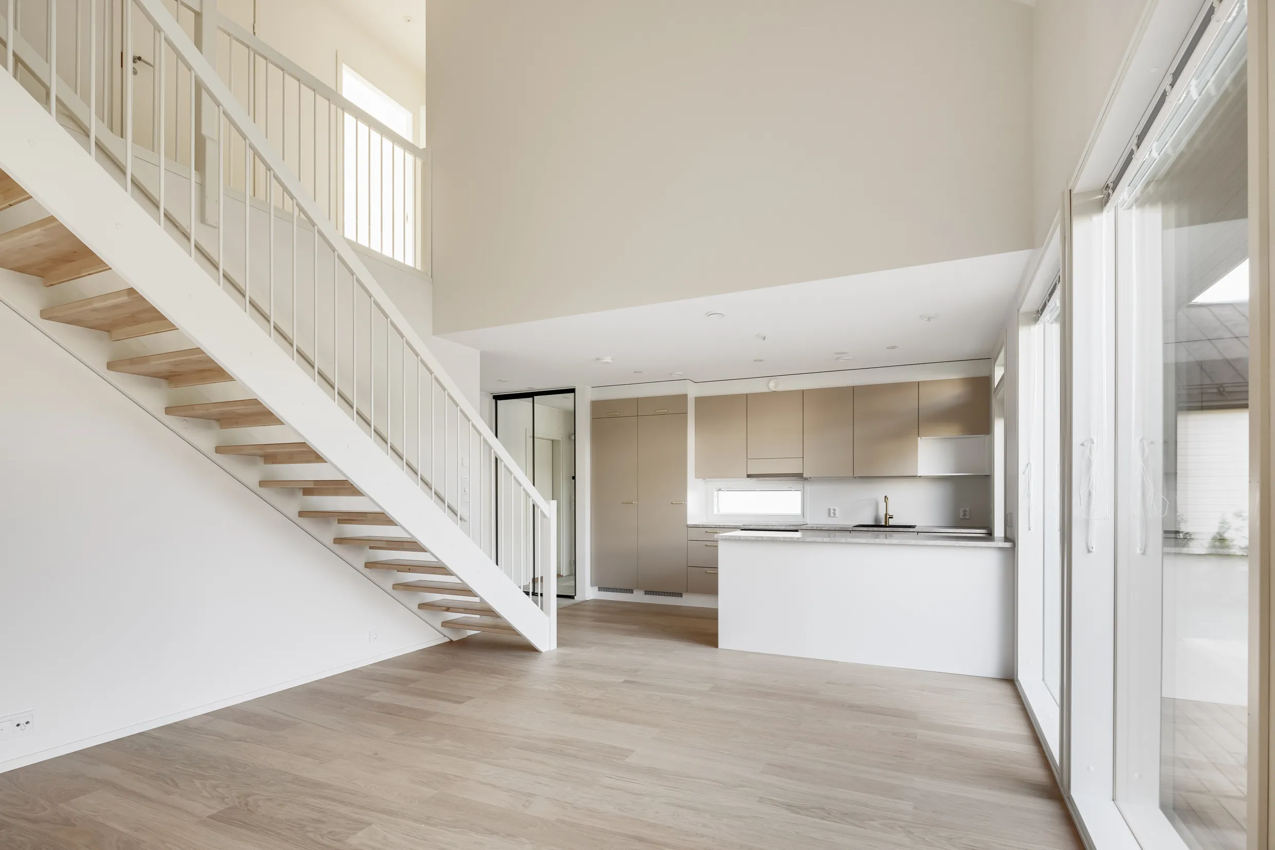 Vaalea, moderni sisätila, jossa on puuportaat, vaaleat keittiökaapit ja suuret ikkunat, jotka tuovat runsaasti luonnonvaloa.