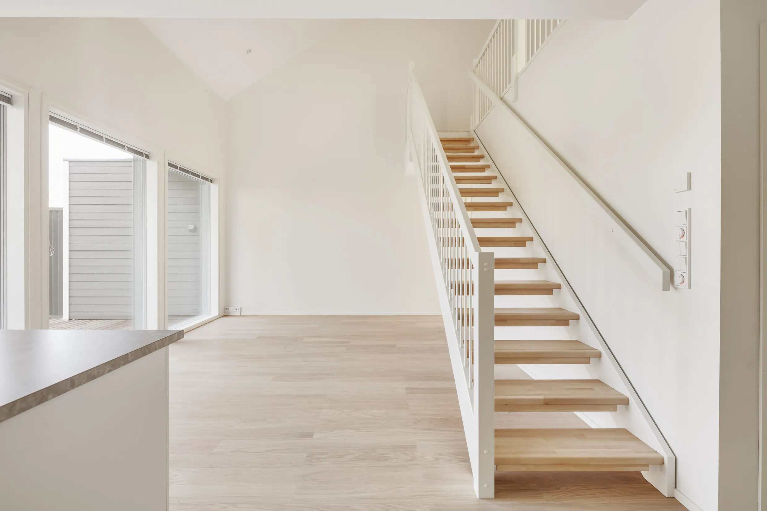 Vaalea, moderni sisätila, jossa puuportaat, suuret ikkunat ja valkoiset seinät luovat avaran ja valoisan tunnelman.