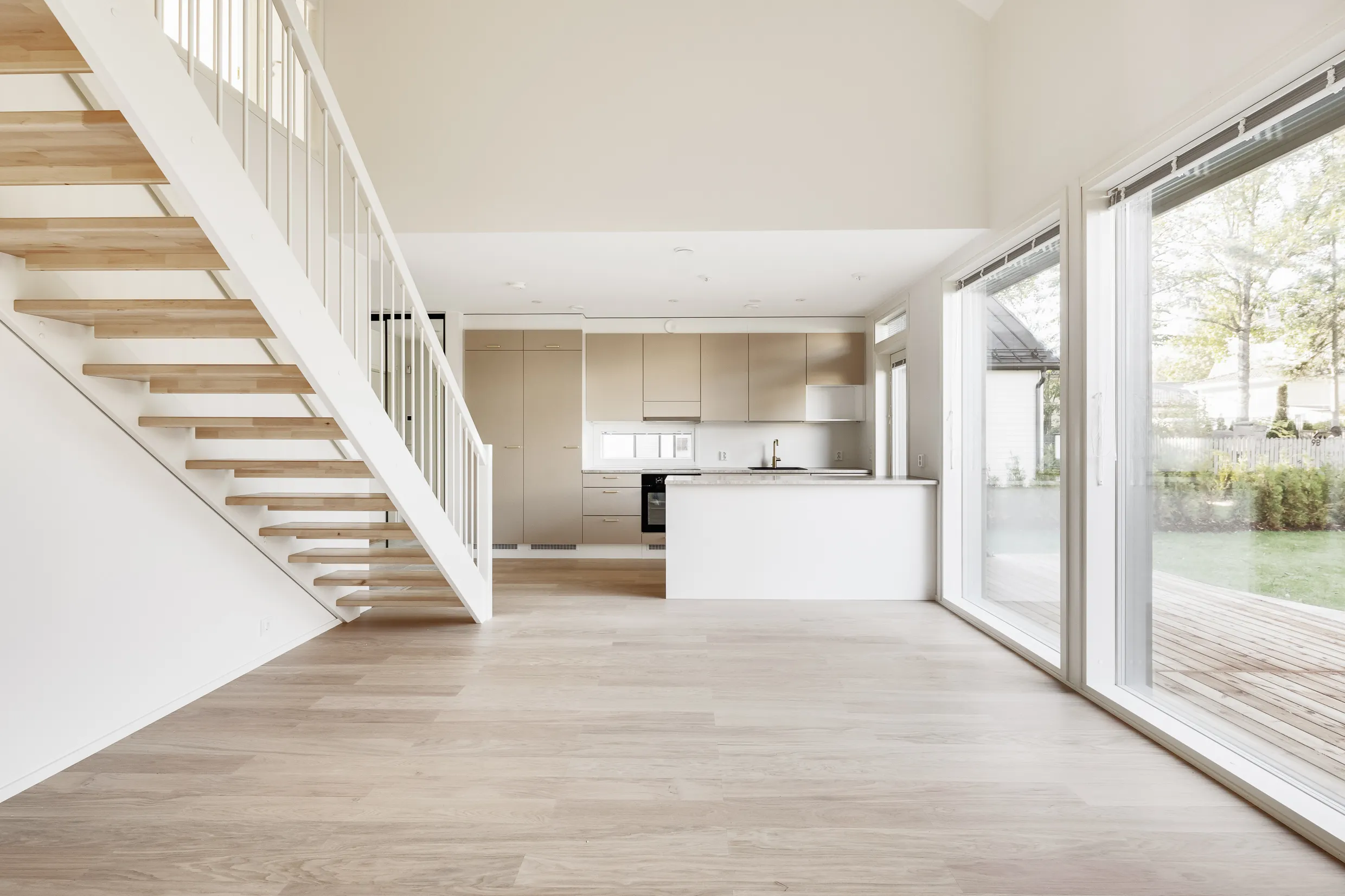 Vaalea ja avara sisätila, jossa on puuportaat, moderni keittiö ja suuret ikkunat, jotka tuovat luonnonvaloa.