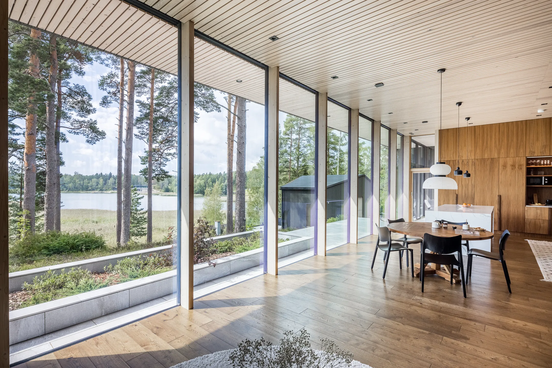 Valoisa sisätila, jossa suuret ikkunat, puulattia, ruokapöytä ja näkymä järvelle.