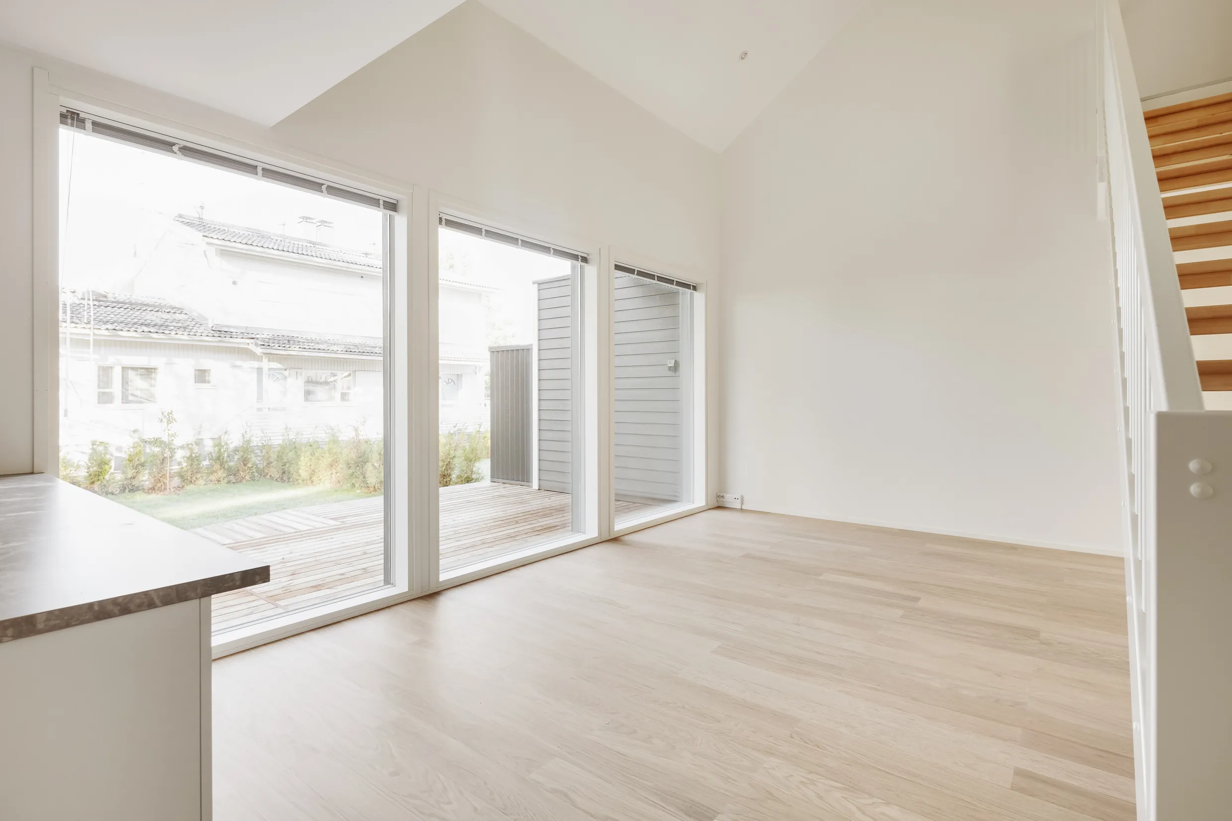 Vaalea, tyhjä huone, jossa on suuret ikkunat, vaaleat puulattiat ja valkoiset seinät.